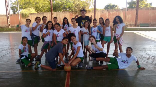 Foto de grupo de jovens em quadra com bola. Grupo de crianças em camisetas brancas e shorts verdes posa em uma quadra, alguns com polegares para cima, com uma bola no centro.