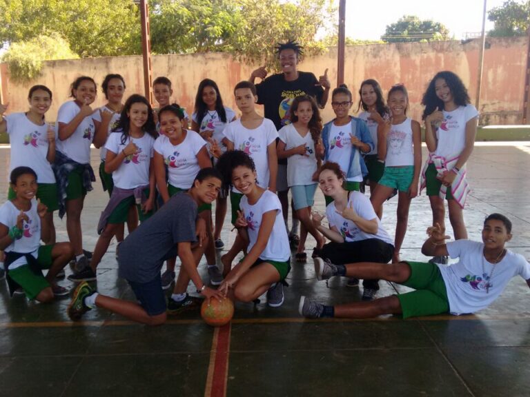 Foto de grupo de jovens em quadra com bola. Grupo de crianças em camisetas brancas e shorts verdes posa em uma quadra, alguns com polegares para cima, com uma bola no centro.