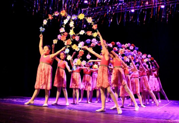 Coreografia floral: apresentação de balé infantil com guirlanda de flores. Dançarinas infantis em vestidos rosa com as mãos erguidas seguram uma guirlanda de flores em uma performance no palco.