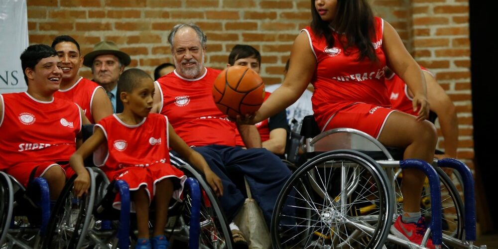 Rafael Cautella Fotografia Jogadores de basquete em cadeiras de rodas, com uniformes vermelhos, interagem com uma bola de basquete em um evento.