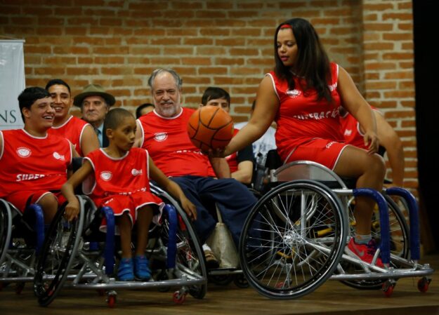 Rafael Cautella Fotografia Jogadores de basquete em cadeiras de rodas, com uniformes vermelhos, interagem com uma bola de basquete em um evento.