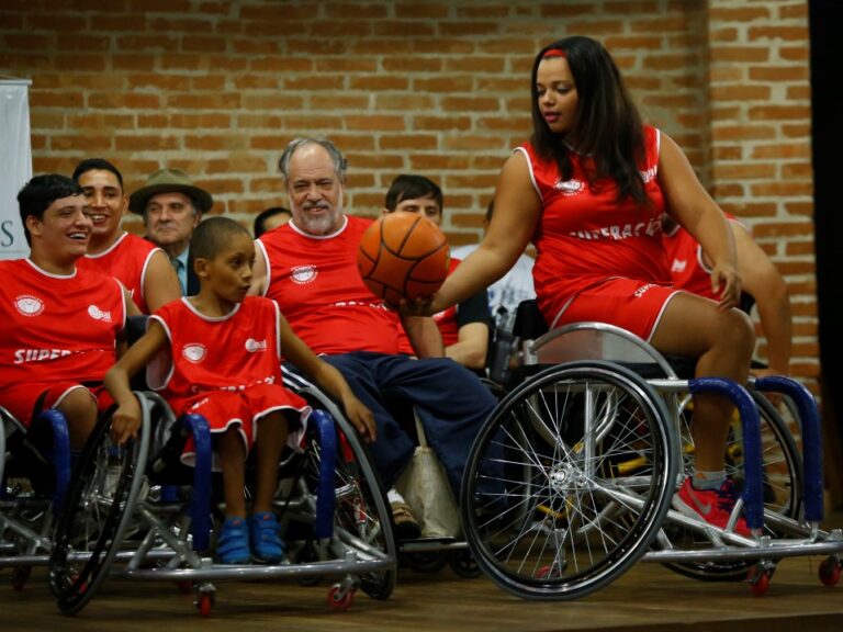 Rafael Cautella Fotografia Jogadores de basquete em cadeiras de rodas, com uniformes vermelhos, interagem com uma bola de basquete em um evento.