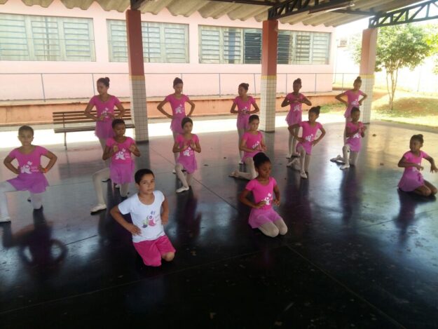 Coreografia de balé infantil em grupo com figurino rosa. Grupo de meninas, a maioria em trajes de balé cor de rosa, em diferentes poses e níveis em uma quadra externa coberta.