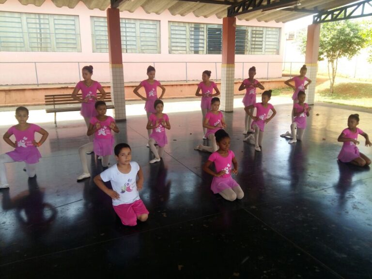 Coreografia de balé infantil em grupo com figurino rosa. Grupo de meninas, a maioria em trajes de balé cor de rosa, em diferentes poses e níveis em uma quadra externa coberta.