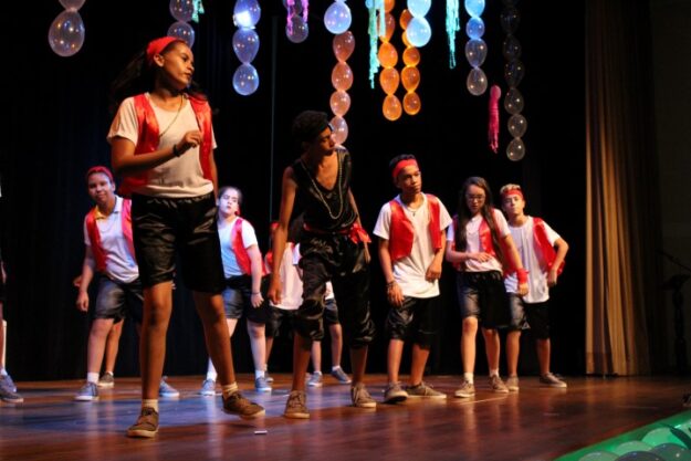 Apresentação de dança jovem em palco. Grupo de jovens dançarinos em trajes brancos, pretos e vermelhos se apresenta no palco, com balões decorando o fundo.