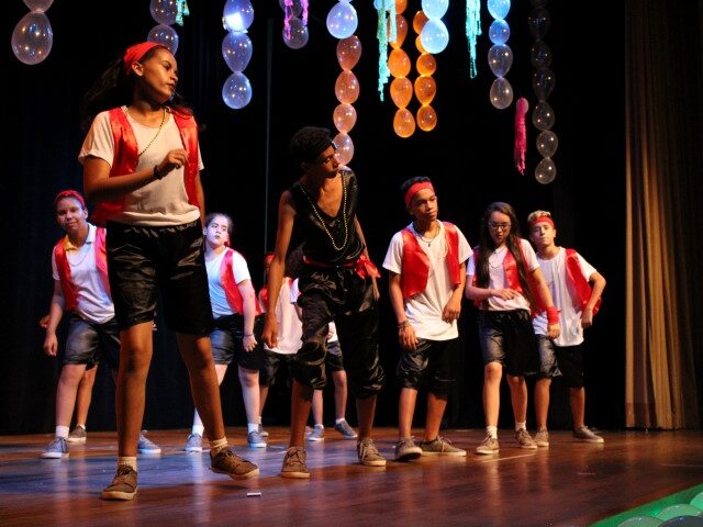 Apresentação de dança jovem em palco. Grupo de jovens dançarinos em trajes brancos, pretos e vermelhos se apresenta no palco, com balões decorando o fundo.