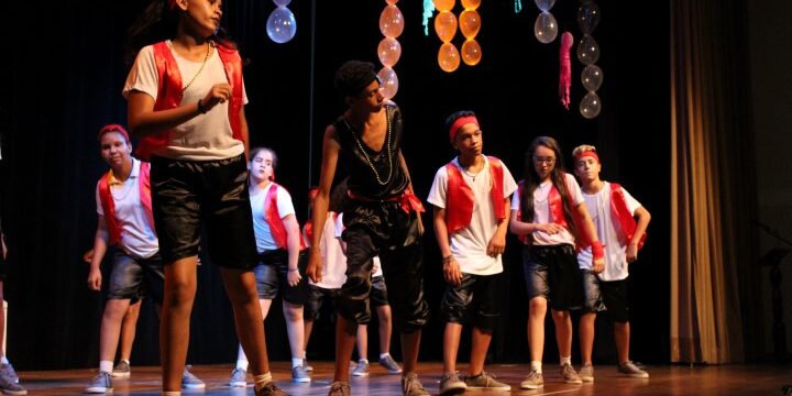 Apresentação de dança jovem em palco. Grupo de jovens dançarinos em trajes brancos, pretos e vermelhos se apresenta no palco, com balões decorando o fundo.