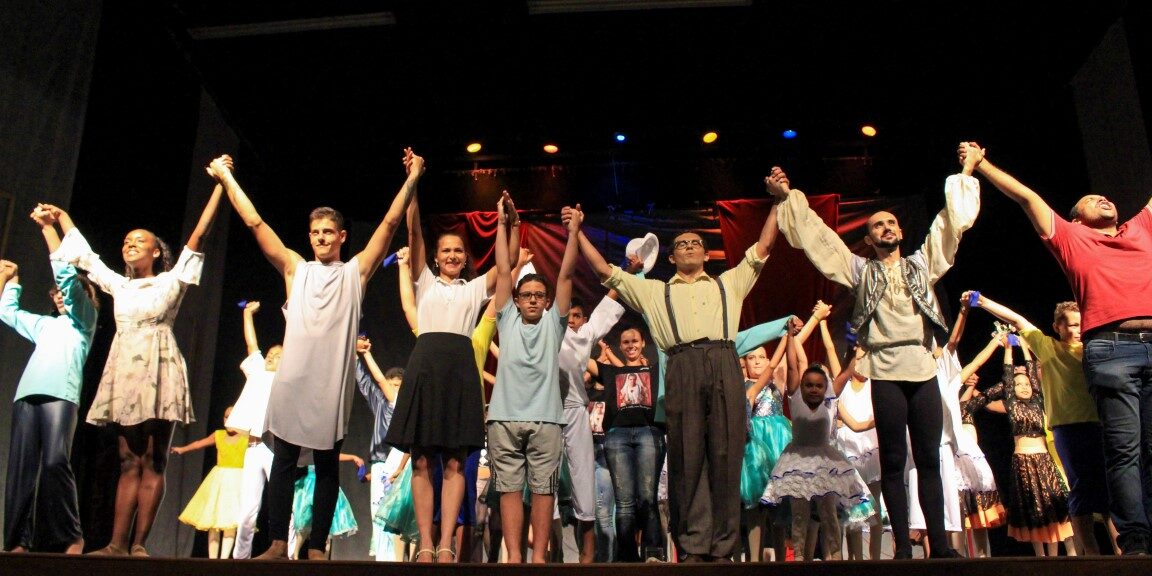 O elenco se curva de mãos dadas em um palco escuro. Elenco de apresentação em grupo, de mãos dadas, com os braços erguidos em um palco escuro para o aplauso final.