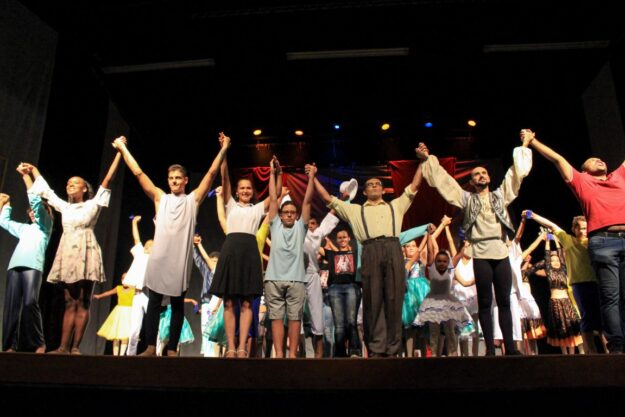 O elenco se curva de mãos dadas em um palco escuro. Elenco de apresentação em grupo, de mãos dadas, com os braços erguidos em um palco escuro para o aplauso final.