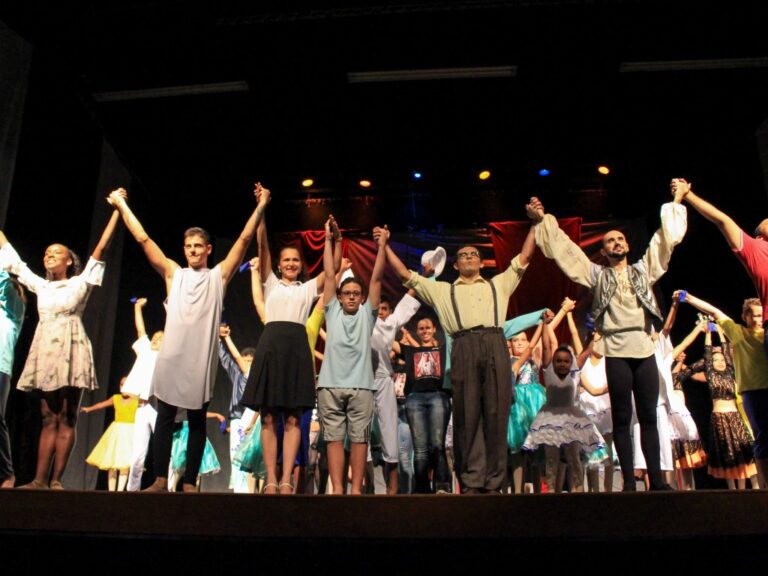 O elenco se curva de mãos dadas em um palco escuro. Elenco de apresentação em grupo, de mãos dadas, com os braços erguidos em um palco escuro para o aplauso final.