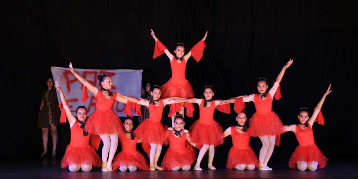 Apresentação de balé em grupo. Grupo de meninas em vestidos vermelhos de balé em uma formação em pirâmide no palco escuro, com os braços estendidos.