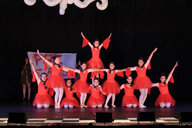 Apresentação de balé em grupo. Grupo de meninas em vestidos vermelhos de balé em uma formação em pirâmide no palco escuro, com os braços estendidos.