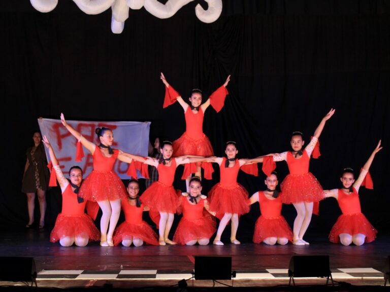 Apresentação de balé em grupo. Grupo de meninas em vestidos vermelhos de balé em uma formação em pirâmide no palco escuro, com os braços estendidos.