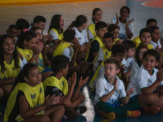 Alunos sentados aplaudem em evento esportivo. Várias crianças e adolescentes, a maioria com coletes amarelos, sentadas no chão de uma quadra, batem palmas e olham para frente.