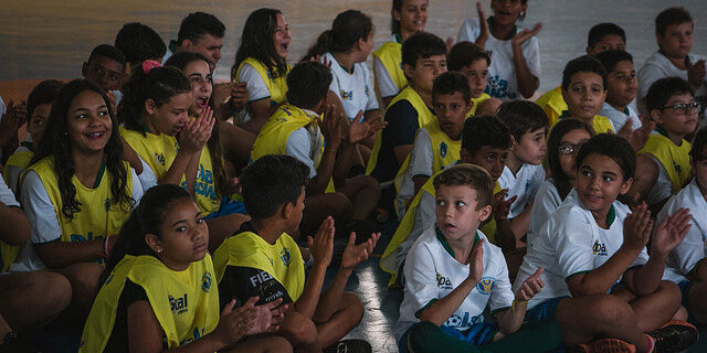 Alunos sentados aplaudem em evento esportivo. Várias crianças e adolescentes, a maioria com coletes amarelos, sentadas no chão de uma quadra, batem palmas e olham para frente.