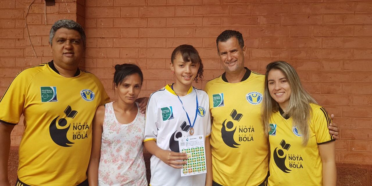 Família e equipe celebram conquista de jovem aluna com medalha e certificado. Cinco pessoas posam para uma foto em frente a uma parede de tijolos, três vestindo camisetas amarelas. A garota no centro segura uma medalha.