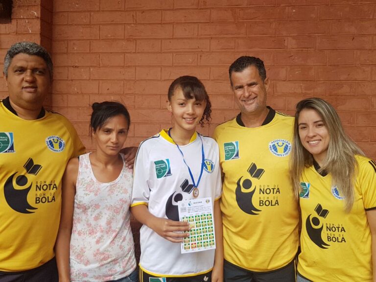 Família e equipe celebram conquista de jovem aluna com medalha e certificado. Cinco pessoas posam para uma foto em frente a uma parede de tijolos, três vestindo camisetas amarelas. A garota no centro segura uma medalha.
