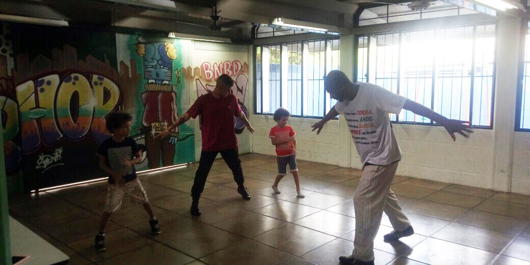 Aula de dança em grupo em sala com grafite. Dois adultos e duas crianças praticam movimentos de dança em uma sala com paredes grafitadas, com luz solar entrando.