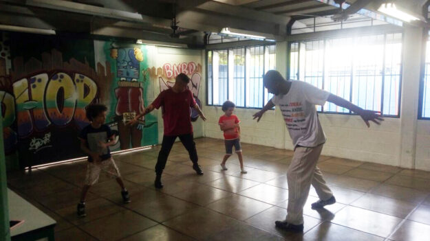 Aula de dança em grupo em sala com grafite. Dois adultos e duas crianças praticam movimentos de dança em uma sala com paredes grafitadas, com luz solar entrando.