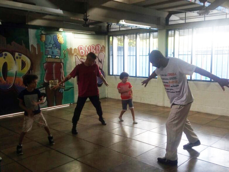 Aula de dança em grupo em sala com grafite. Dois adultos e duas crianças praticam movimentos de dança em uma sala com paredes grafitadas, com luz solar entrando.