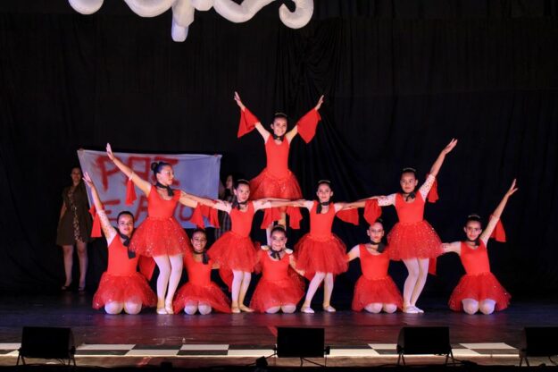 Apresentação de balé em grupo. Grupo de meninas em vestidos vermelhos de balé em uma formação em pirâmide no palco escuro, com os braços estendidos.
