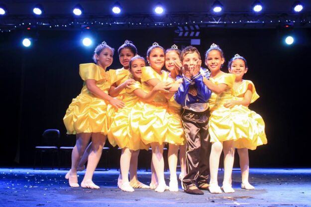Dançarinos infantis com trajes de princesa e príncipe no palco. Um grupo de sete meninas de amarelo e um menino de azul e marrom, em trajes de dança, posam no palco.