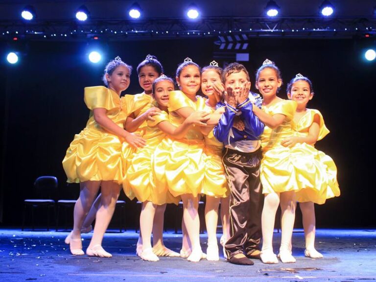 Dançarinos infantis com trajes de princesa e príncipe no palco. Um grupo de sete meninas de amarelo e um menino de azul e marrom, em trajes de dança, posam no palco.