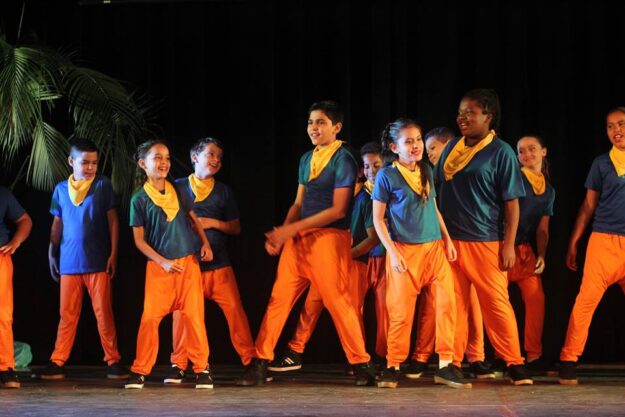 Crianças em trajes de dança coloridos se apresentam. Grupo de crianças e adolescentes com calças laranjas e camisetas azuis com lenços amarelos se apresenta no palco.