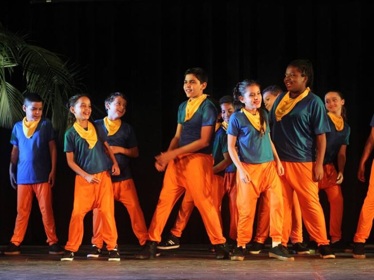 Crianças em trajes de dança coloridos se apresentam. Grupo de crianças e adolescentes com calças laranjas e camisetas azuis com lenços amarelos se apresenta no palco.