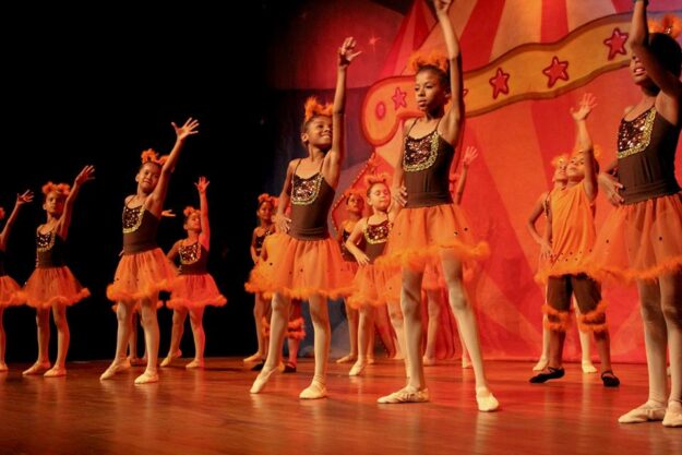 Apresentação de balé infantil com tema circense. Meninas em trajes de balé marrons e laranjas com cabelos adornados se apresentam em um palco com um cenário circense.