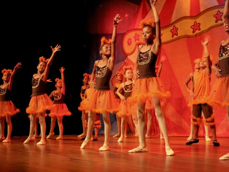 Apresentação de balé infantil com tema circense. Meninas em trajes de balé marrons e laranjas com cabelos adornados se apresentam em um palco com um cenário circense.