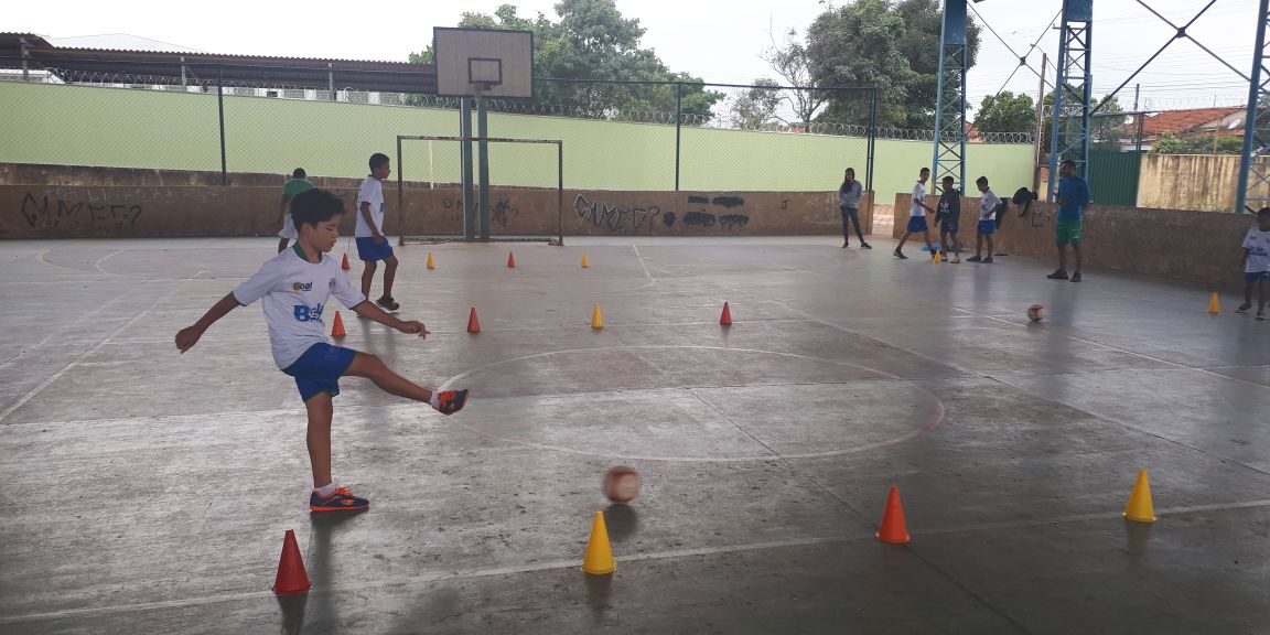 Treino de futebol com drible em quadra. Jovem em quadra poliesportiva coberta chuta uma bola de futebol durante um exercício de drible com cones laranja e amarelos.