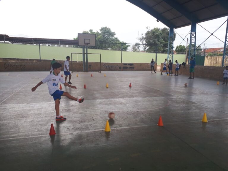 Treino de futebol com drible em quadra. Jovem em quadra poliesportiva coberta chuta uma bola de futebol durante um exercício de drible com cones laranja e amarelos.