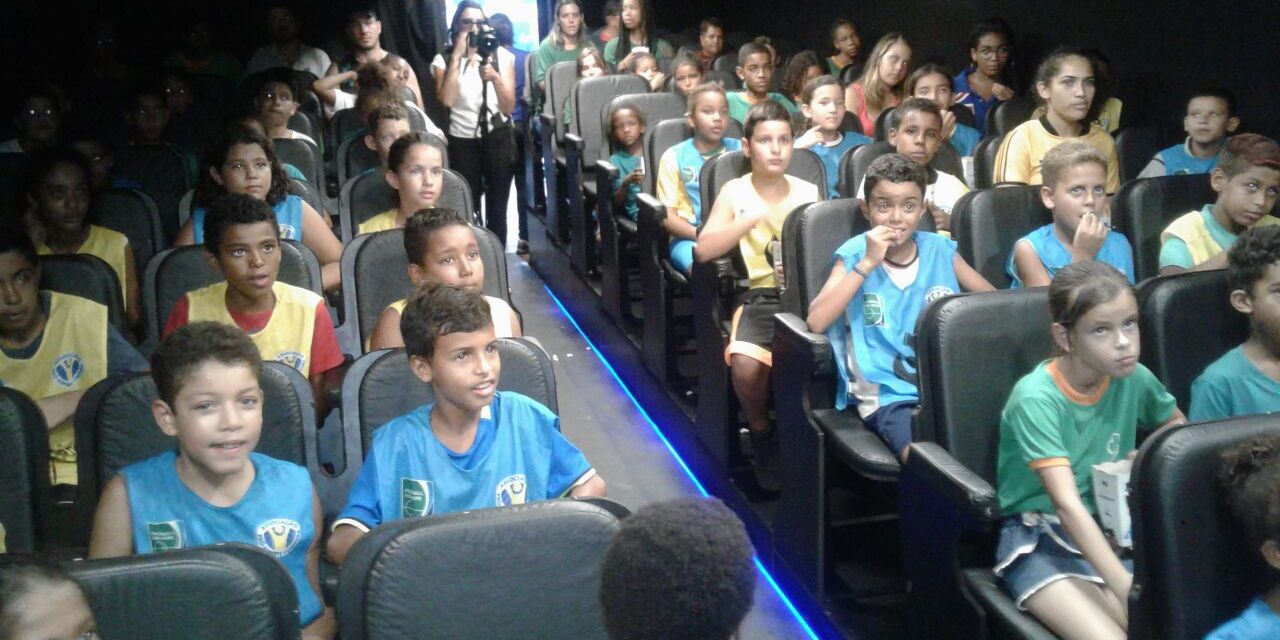 Plateia infantil assiste a uma exibição em auditório. Várias crianças em uniformes esportivos coloridos estão sentadas em assentos pretos de um auditório, olhando para frente.