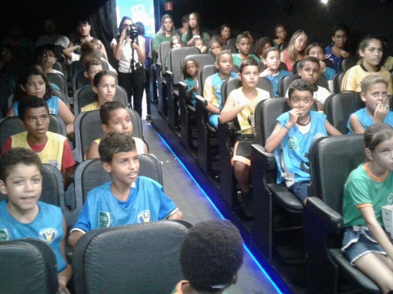 Plateia infantil assiste a uma exibição em auditório. Várias crianças em uniformes esportivos coloridos estão sentadas em assentos pretos de um auditório, olhando para frente.