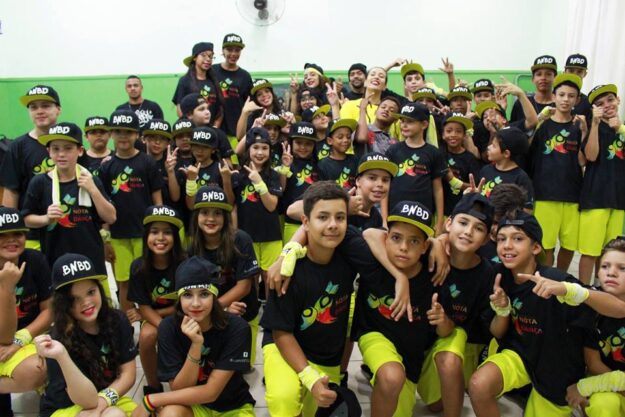 Foto em grupo de jovens dançarinos uniformizados. Grande grupo de crianças e adolescentes com bonés e uniformes pretos e amarelos posa com gestos de alegria para foto.
