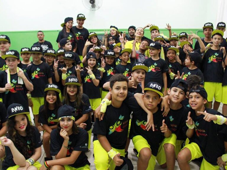 Foto em grupo de jovens dançarinos uniformizados. Grande grupo de crianças e adolescentes com bonés e uniformes pretos e amarelos posa com gestos de alegria para foto.