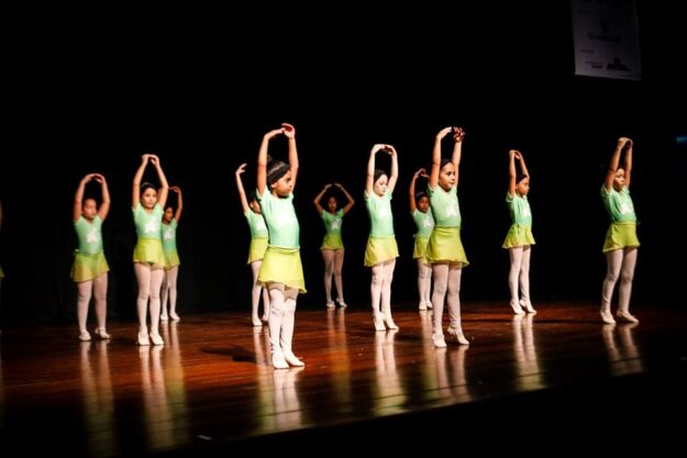 Apresentação de balé infantil em palco. Meninas em uniformes de balé verde-claro e meias brancas se apresentam em um palco escuro de madeira com as mãos erguidas.