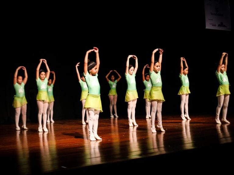 Apresentação de balé infantil em palco. Meninas em uniformes de balé verde-claro e meias brancas se apresentam em um palco escuro de madeira com as mãos erguidas.