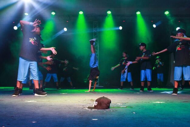 Performance de jovens no palco. Grupo de jovens se apresenta no palco com um dançarino de breakdance fazendo um headstand sob luzes verdes.