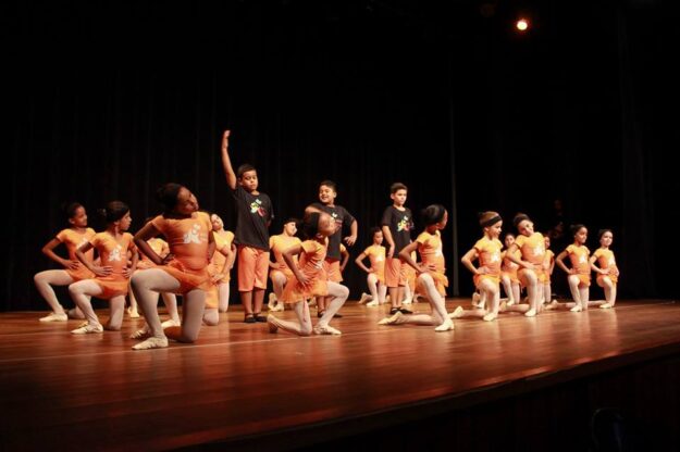Apresentação de dança infantil no palco. Crianças em uniformes de dança laranja e meias brancas se apresentam no palco escuro, com alguns em joelhos e outros em pé.