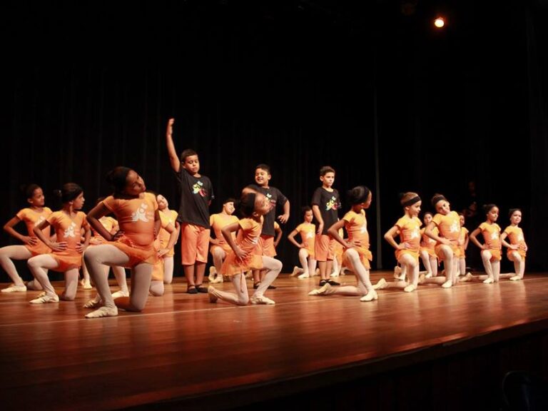 Apresentação de dança infantil no palco. Crianças em uniformes de dança laranja e meias brancas se apresentam no palco escuro, com alguns em joelhos e outros em pé.