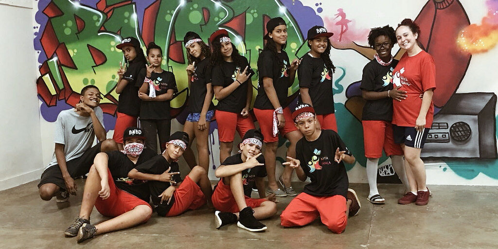 Grupo de dança em pose descontraída com fundo de grafite. Grupo de jovens, a maioria em camisetas pretas e shorts vermelhos, posa em frente a uma parede com grafite colorido.