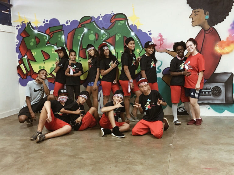 Grupo de dança em pose descontraída com fundo de grafite. Grupo de jovens, a maioria em camisetas pretas e shorts vermelhos, posa em frente a uma parede com grafite colorido.