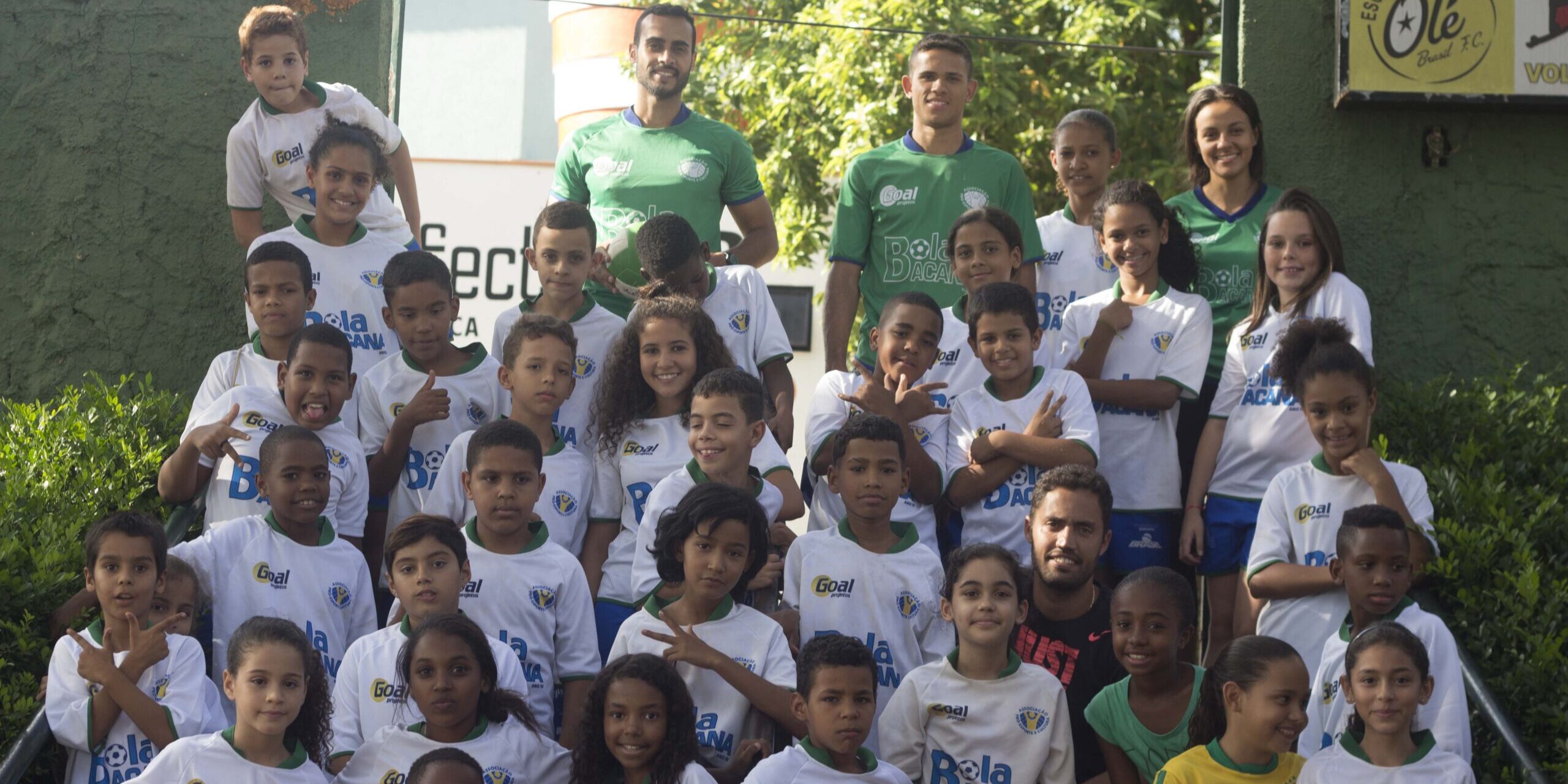 Equipe de futebol e alunos. Grande grupo de crianças e adultos em uniformes de futebol, posando para foto em escadaria externa com folhagens.