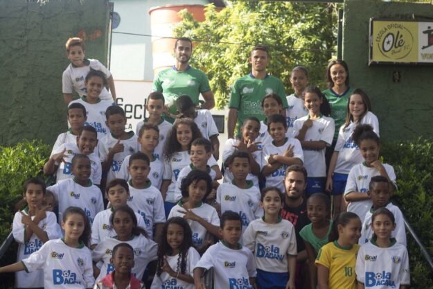 Equipe de futebol e alunos. Grande grupo de crianças e adultos em uniformes de futebol, posando para foto em escadaria externa com folhagens.