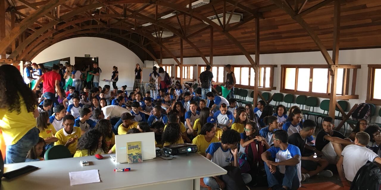 Evento com estudantes em auditório. Estudantes sentados no chão e em cadeiras assistem a uma apresentação com projetor em um auditório com teto de madeira.