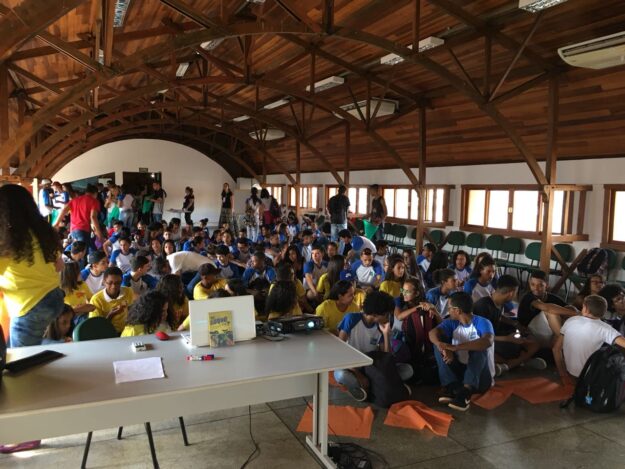 Evento com estudantes em auditório. Estudantes sentados no chão e em cadeiras assistem a uma apresentação com projetor em um auditório com teto de madeira.