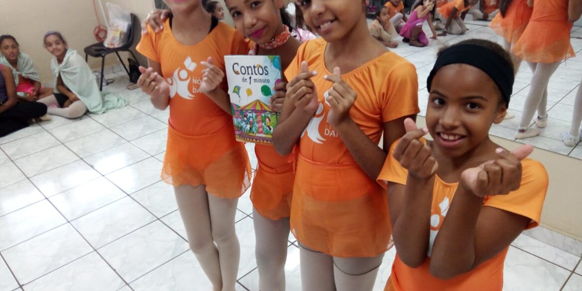 Dança, leitura e gestos de carinho. Quatro crianças bailarinas em collants laranja e meia-calça, sorrindo e fazendo corações com as mãos. Uma delas segura um livro.