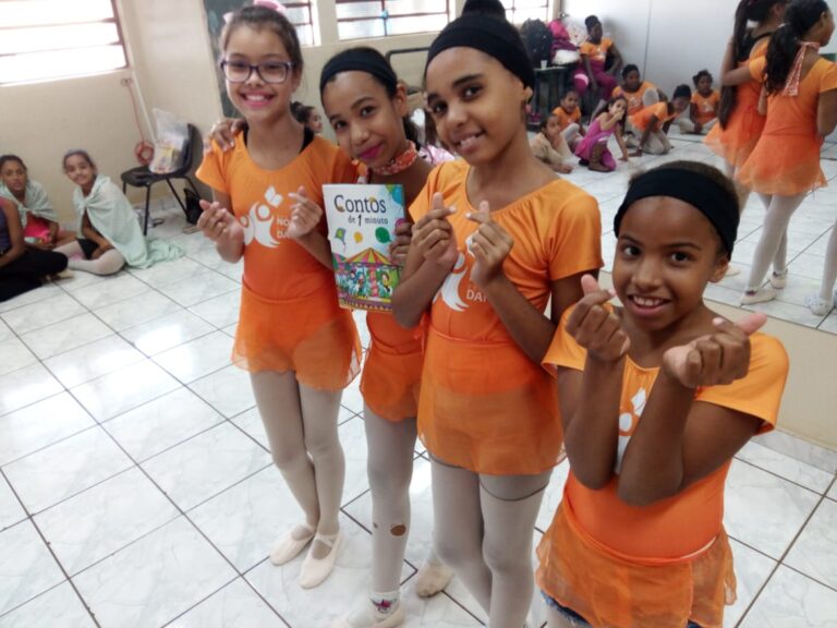 Dança, leitura e gestos de carinho. Quatro crianças bailarinas em collants laranja e meia-calça, sorrindo e fazendo corações com as mãos. Uma delas segura um livro.